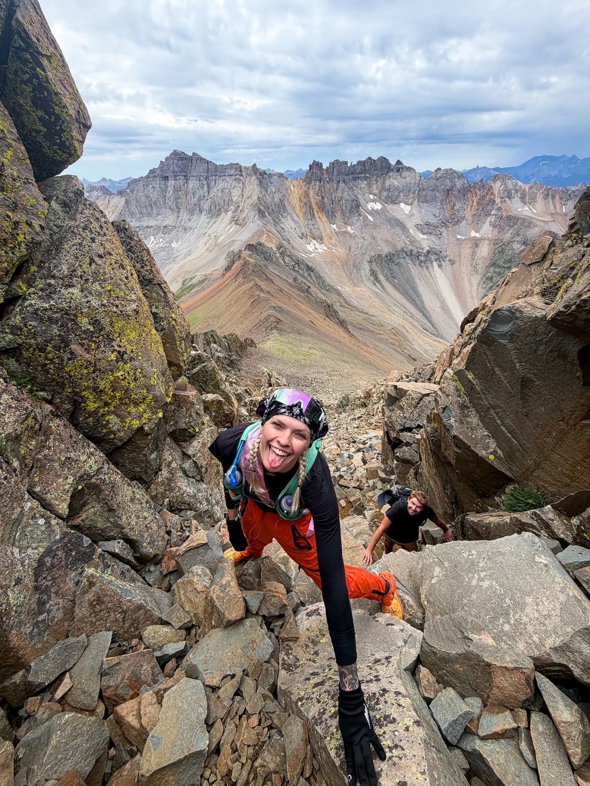 Bri scrambling up a rocky mountain ridge with mountain views behind her